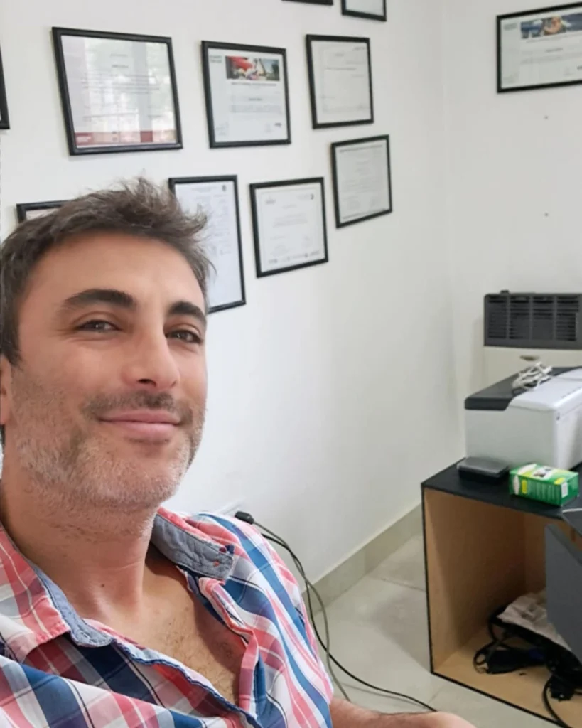 Nutricionista sonriente toma una selfie en su consultorio. En la pared se exhiben múltiples diplomas enmarcados y a un lado se observa una impresora sobre un escritorio.